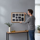 A man is hanging a picture frame on a gray wall, with a wooden table and a vase of greenery nearby.